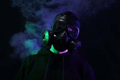 Man wearing gas mask in color lights and smoke on black background, low angle view