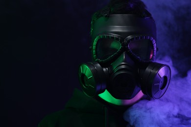 Man wearing gas mask in color lights and smoke on black background. Space for text