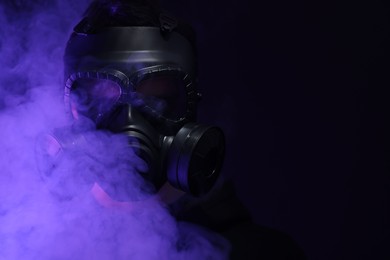 Photo of Man wearing gas mask in color lights and smoke on black background. Space for text