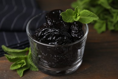 Photo of Tasty dried plums (prunes) in glass bowl and mint leaves on wooden table, closeup