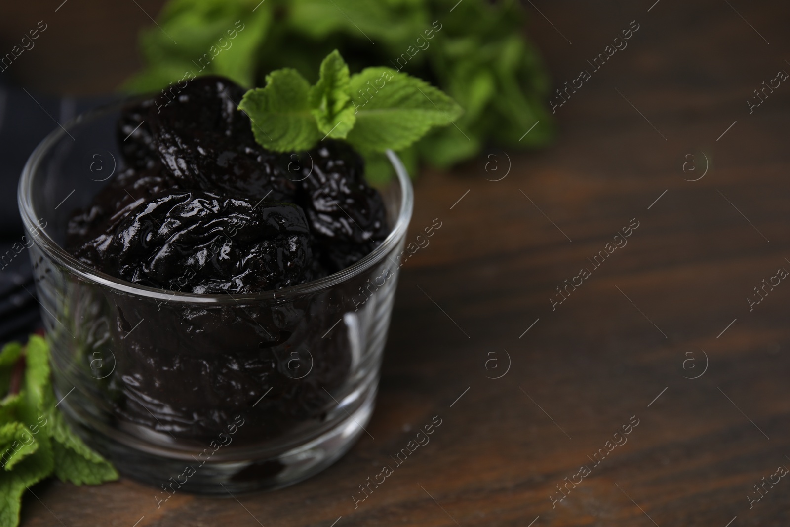 Photo of Tasty dried plums (prunes) in glass bowl and mint leaves on wooden table, closeup. Space for text