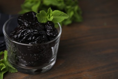 Tasty dried plums (prunes) in glass bowl and mint leaves on wooden table, closeup. Space for text