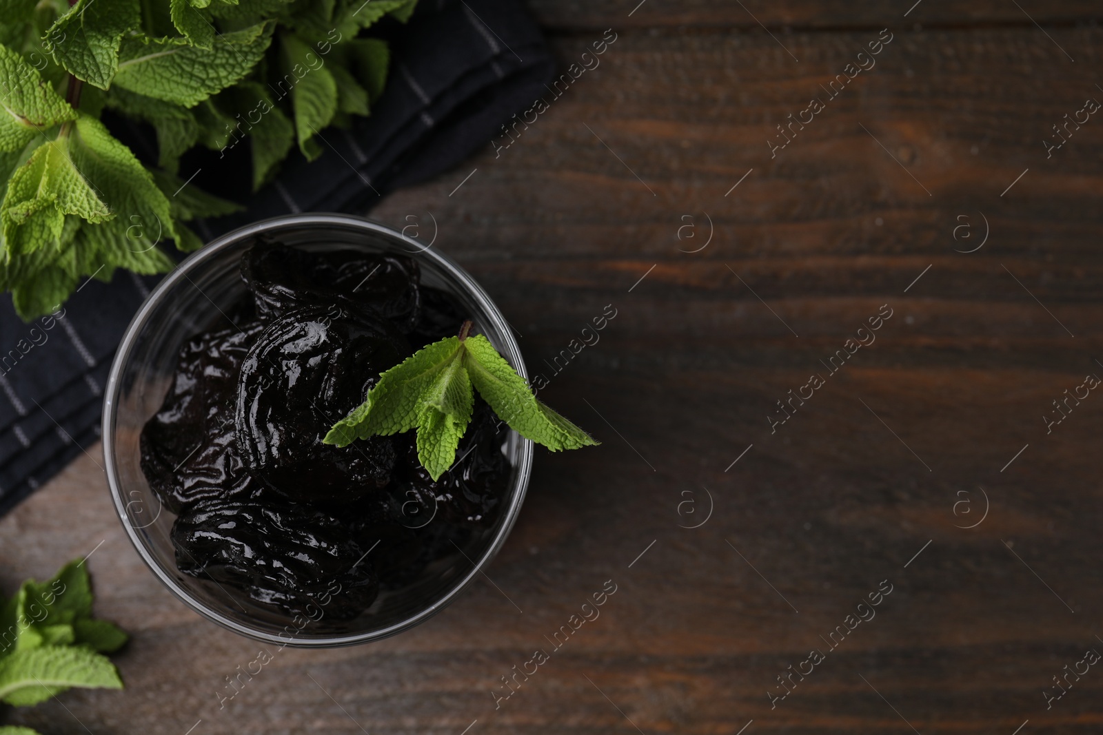 Photo of Tasty dried plums (prunes) in glass bowl and mint leaves on wooden table, flat lay. Space for text