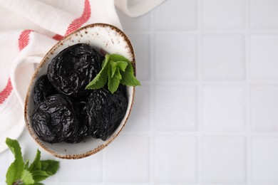 Tasty dried plums (prunes) and mint leaves on white tiled table, flat lay. Space for text