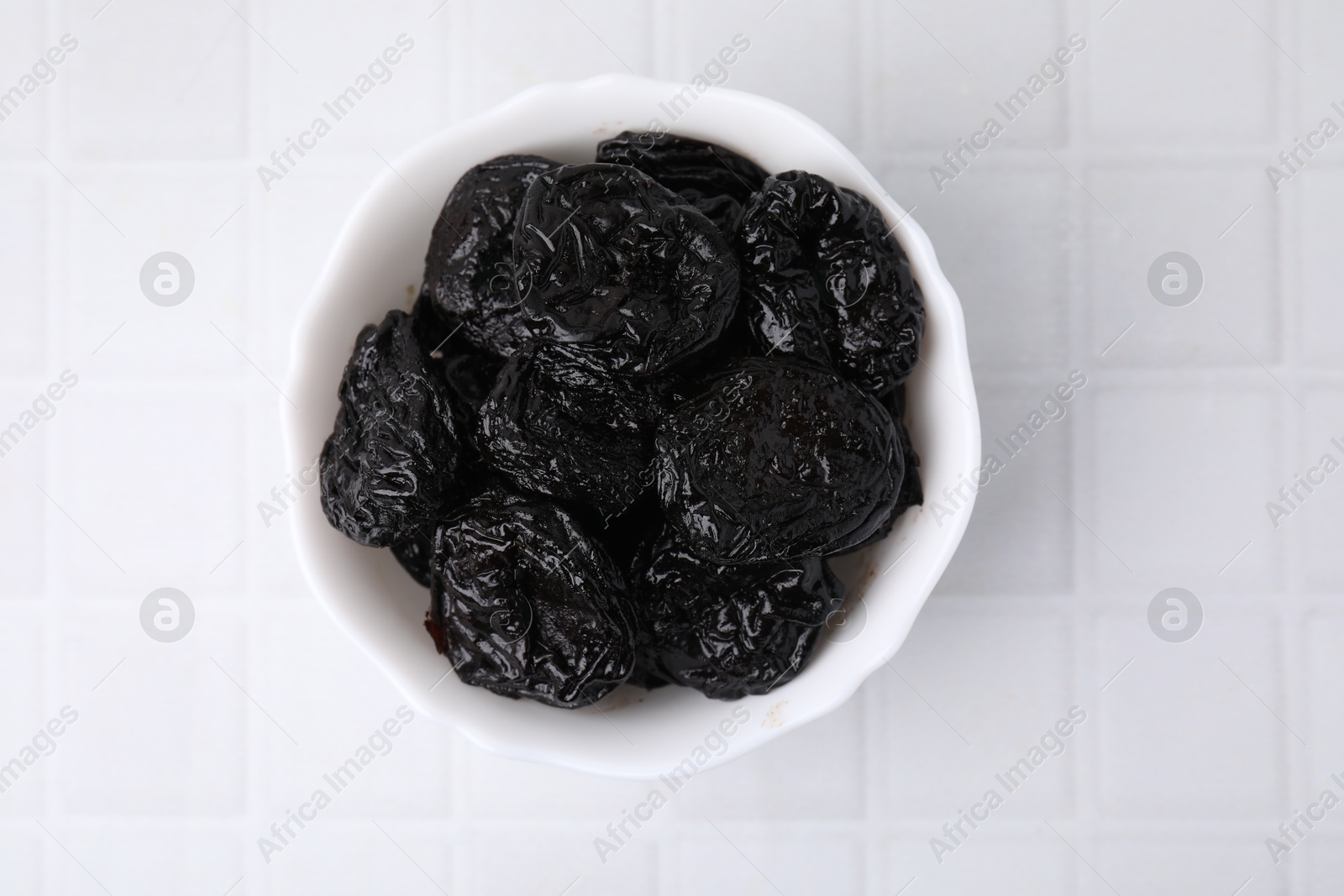 Photo of Tasty dried plums (prunes) in bowl on white tiled table, top view