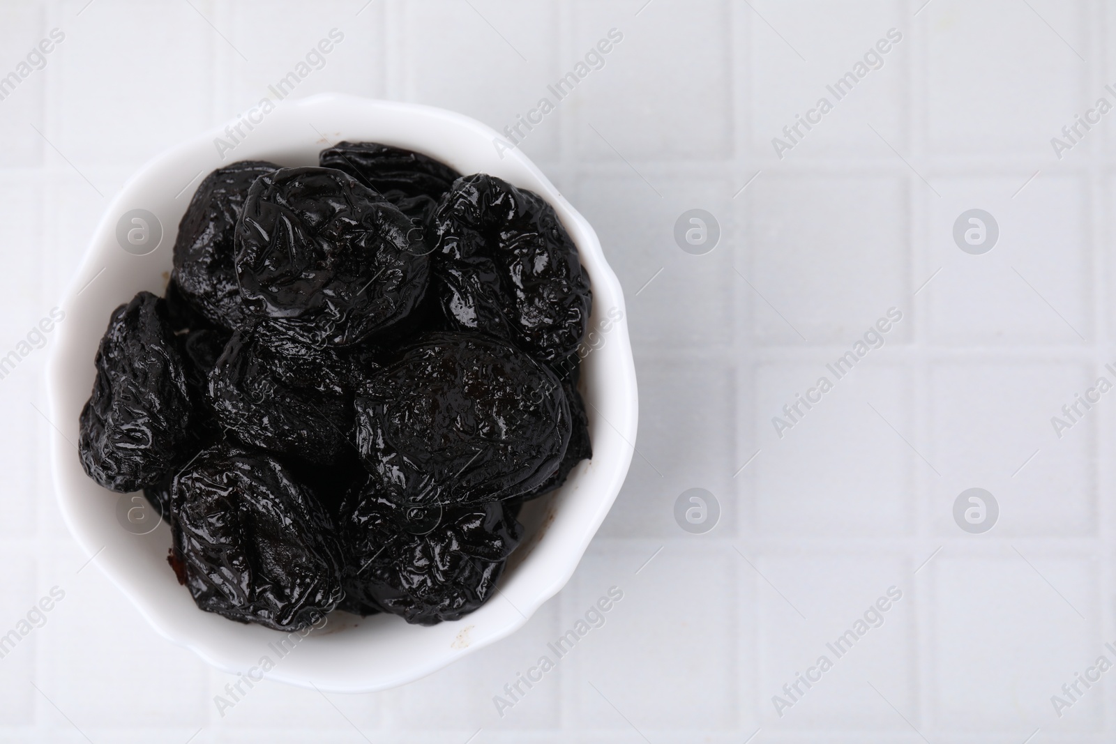 Photo of Tasty dried plums (prunes) in bowl on white tiled table, top view. Space for text