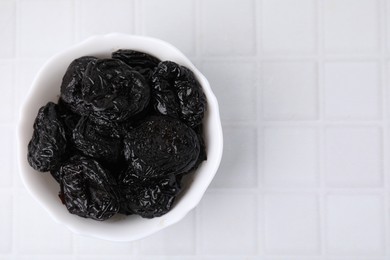 Photo of Tasty dried plums (prunes) in bowl on white tiled table, top view. Space for text