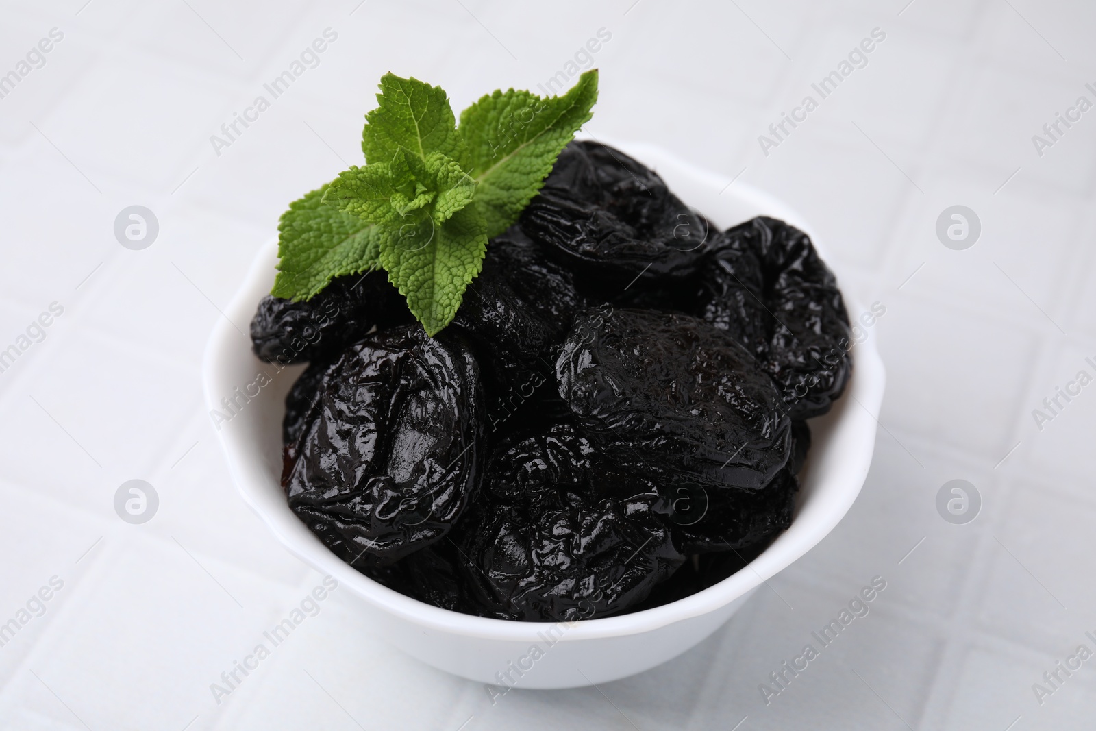 Photo of Tasty dried plums (prunes) and mint leaves on white tiled table, closeup