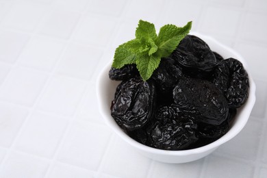 Photo of Tasty dried plums (prunes) and mint leaves on white tiled table, closeup. Space for text