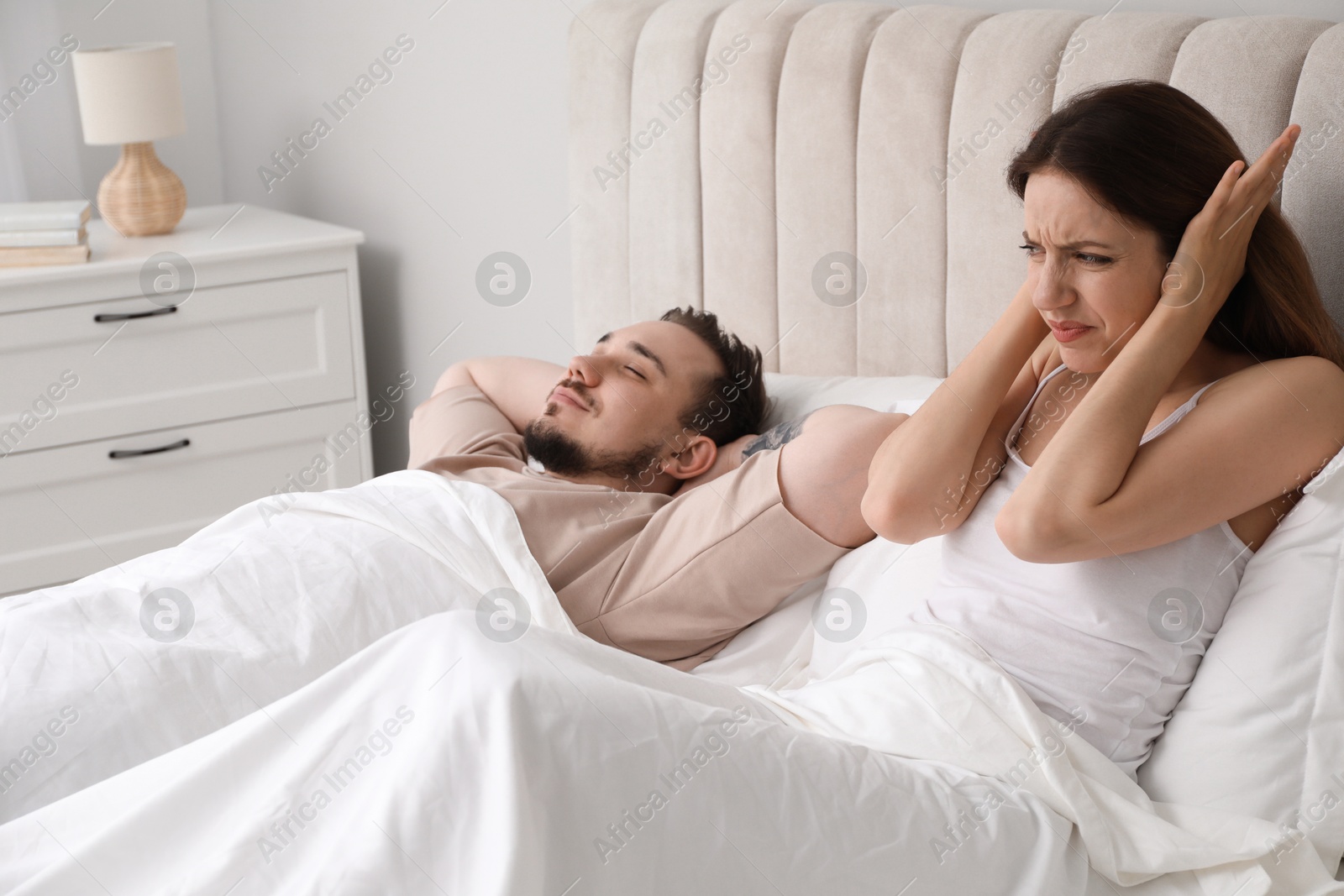 Photo of Bedtime. Irritated woman covering ears near her snoring husband in bed at home