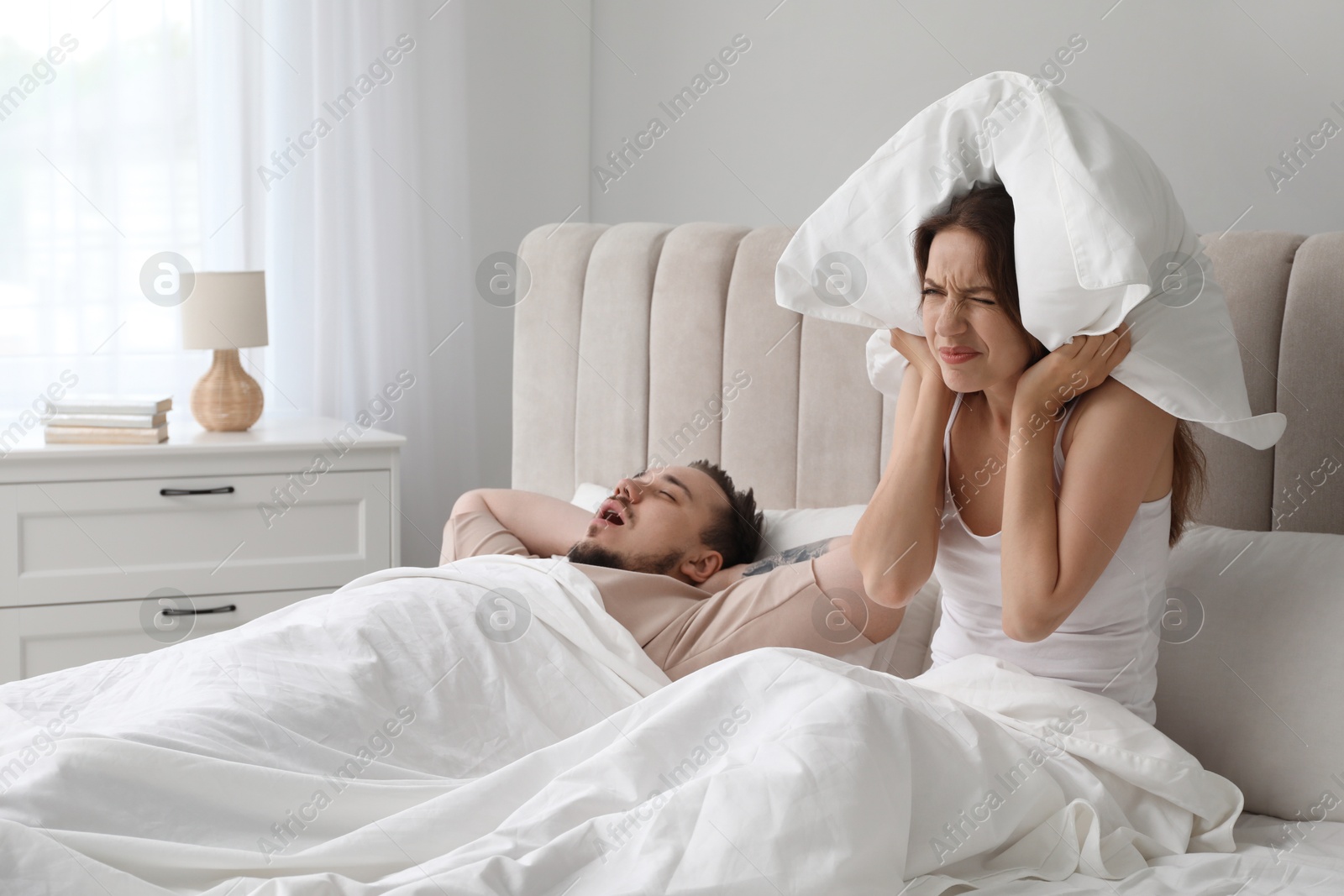 Photo of Bedtime. Irritated woman covering ears near her snoring husband in bed at home