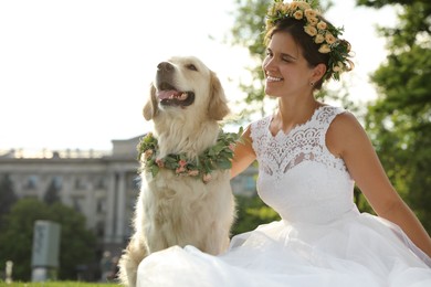 Photo of Bride and adorable Golden Retriever wearing wreath made of beautiful flowers on green grass outdoors