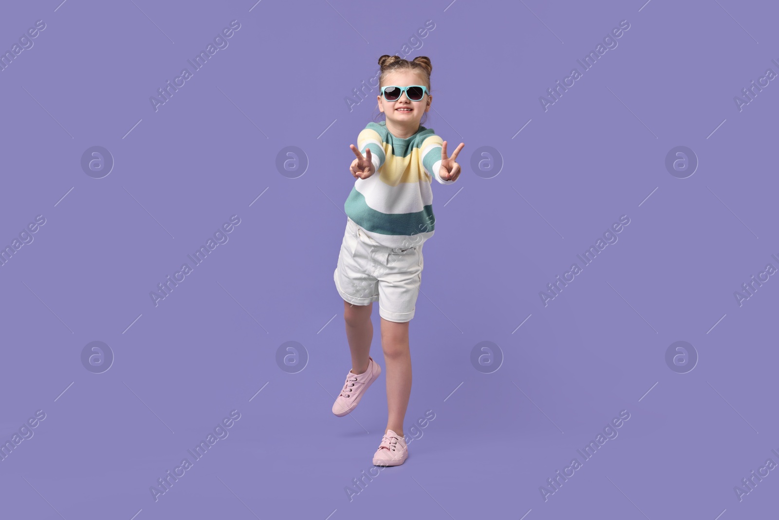 Photo of Cute little girl in sunglasses showing V-sign while dancing on violet background