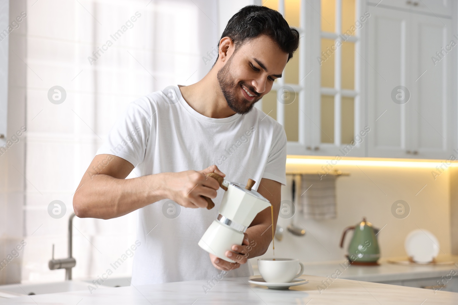Photo of Morning of happy man pouring coffee from moka pot into cup at table in kitchen