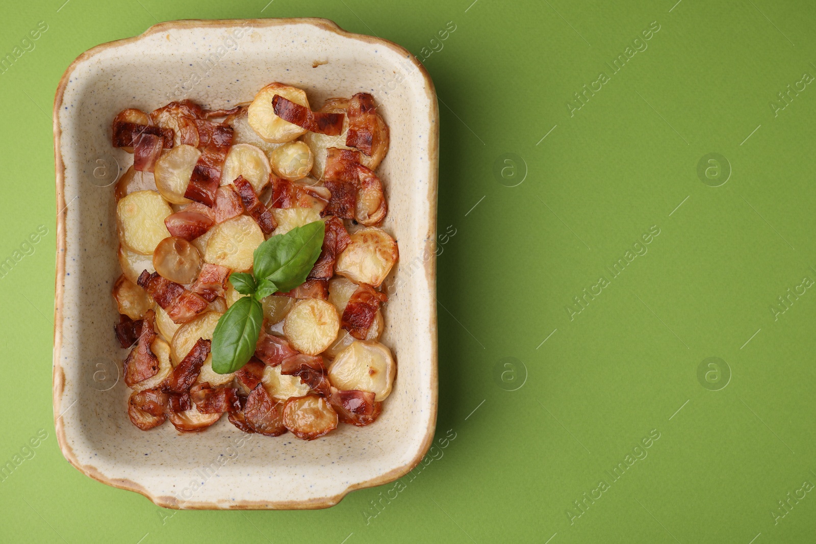 Photo of Delicious baked potatoes, bacon and basil in baking dish on green background, top view. Space for text