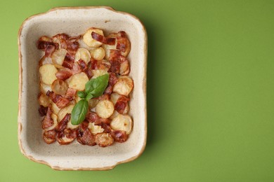 Delicious baked potatoes, bacon and basil in baking dish on green background, top view. Space for text