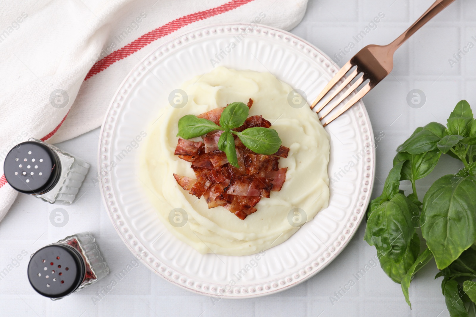 Photo of Fried bacon, mashed potato, basil and fork on white tiled table, top view