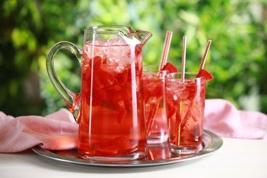 Tasty strawberry lemonade in jug and glasses on white table against blurred green background