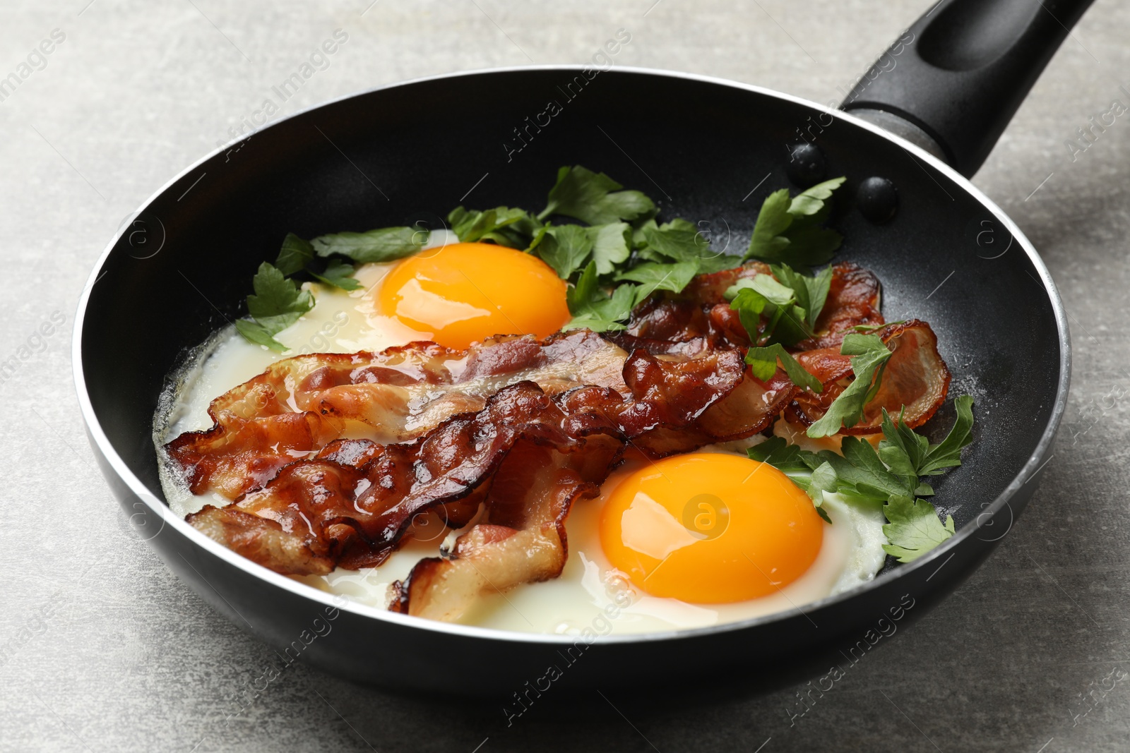 Photo of Tasty bacon, eggs and parsley in frying pan on gray textured table, closeup