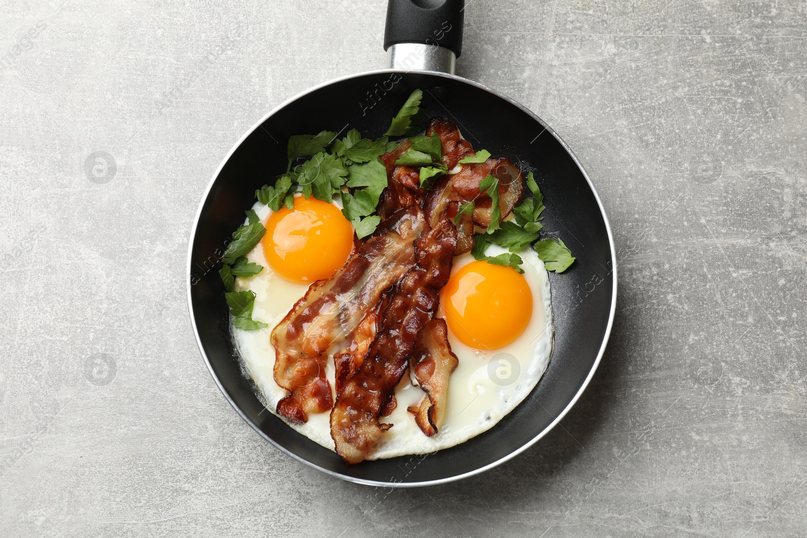 Photo of Tasty bacon, eggs and parsley in frying pan on gray textured table, top view