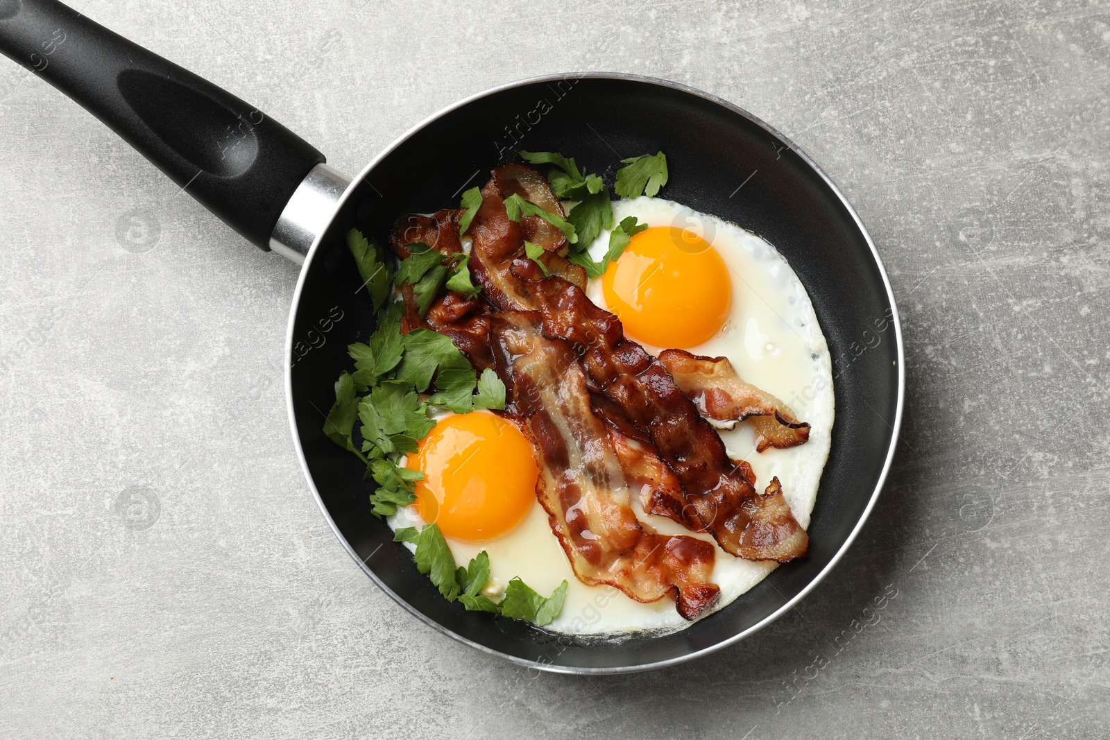 Photo of Tasty bacon, eggs and parsley in frying pan on gray textured table, top view