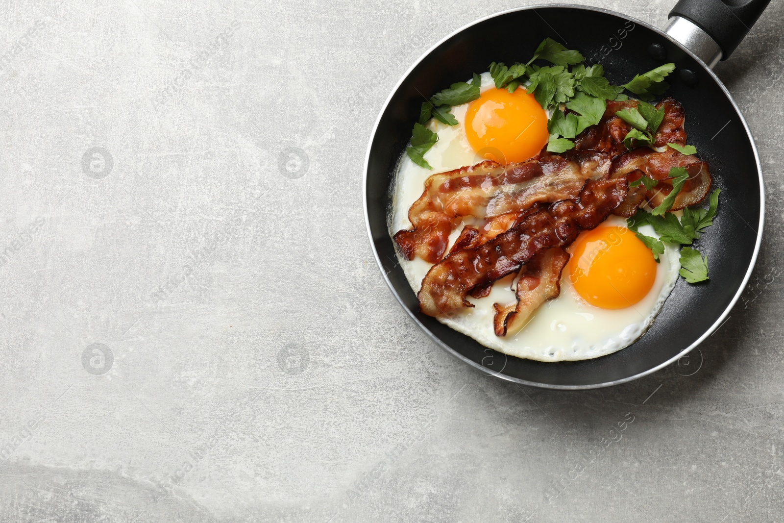 Photo of Tasty bacon, eggs and parsley in frying pan on gray textured table, top view. Space for text