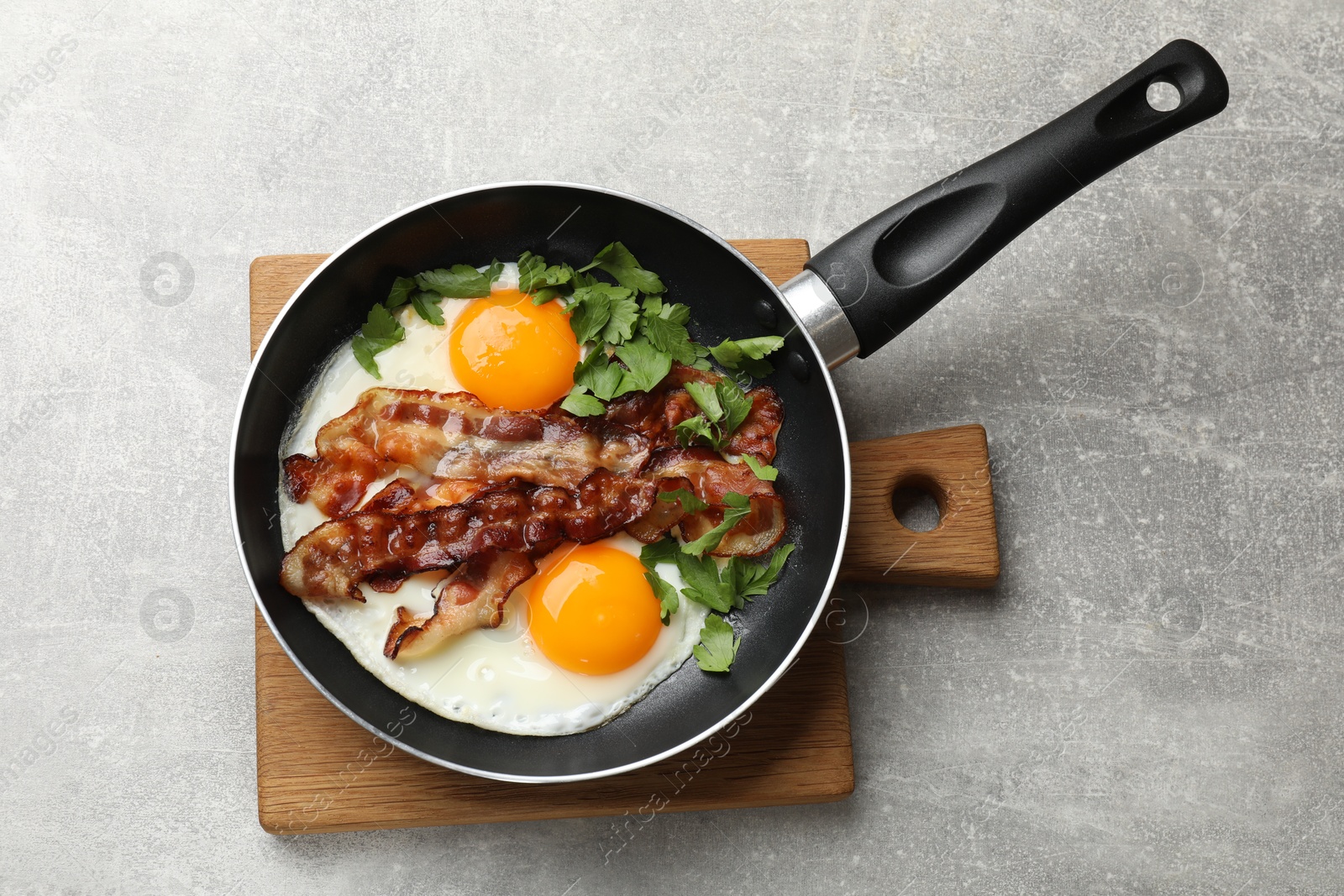 Photo of Tasty bacon, eggs and parsley in frying pan on gray textured table, top view