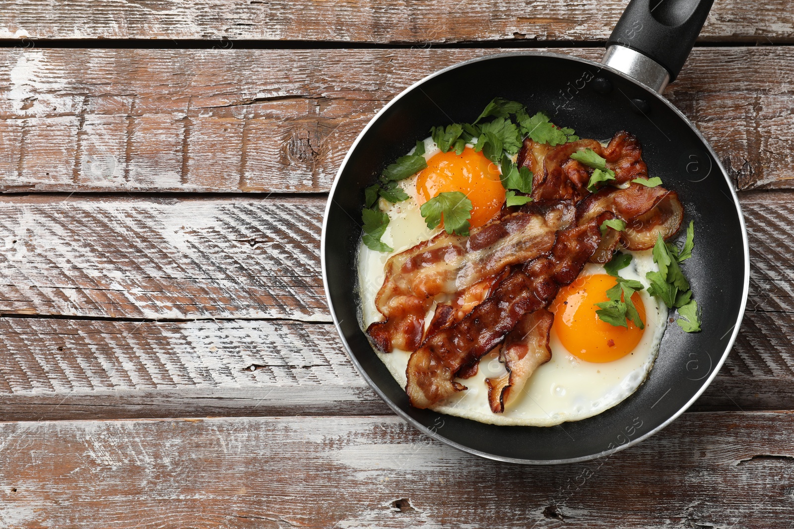 Photo of Tasty bacon, eggs and parsley in frying pan on wooden table, top view. Space for text