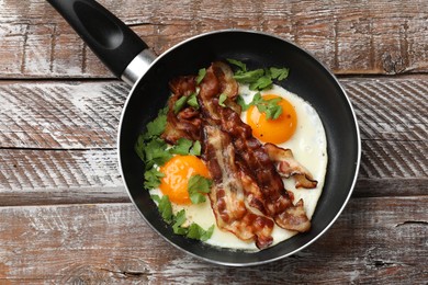 Tasty bacon, eggs and parsley in frying pan on wooden table, top view