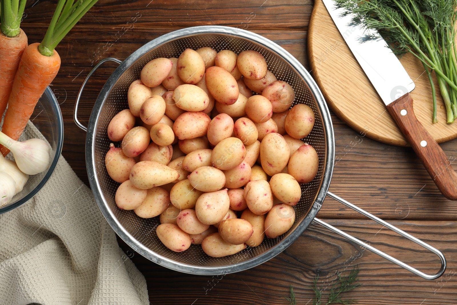 Photo of Raw potatoes in colander, carrots, green onion, dill and knife on wooden table, top view