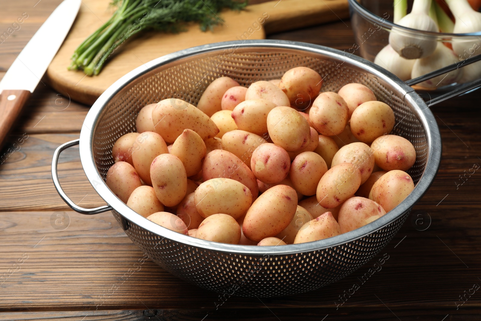 Photo of Raw potatoes in colander, green onion and knife on wooden table