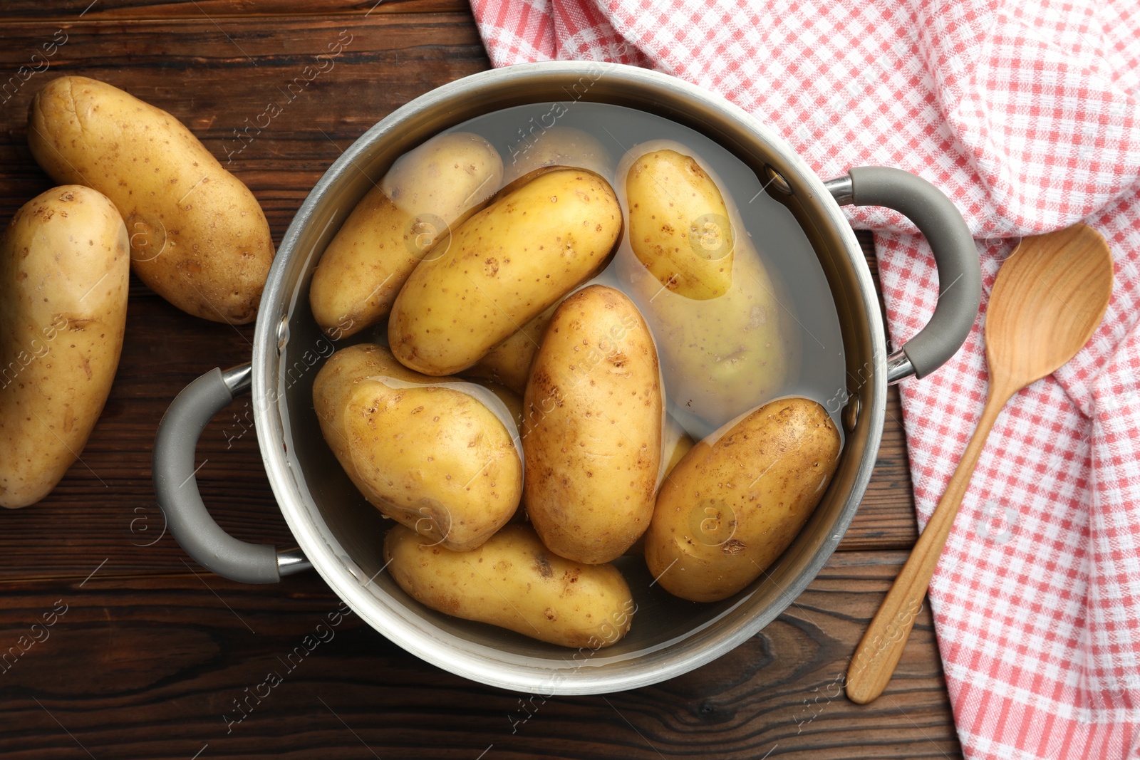 Photo of Raw potatoes in pot with water and spoon on wooden table, top view