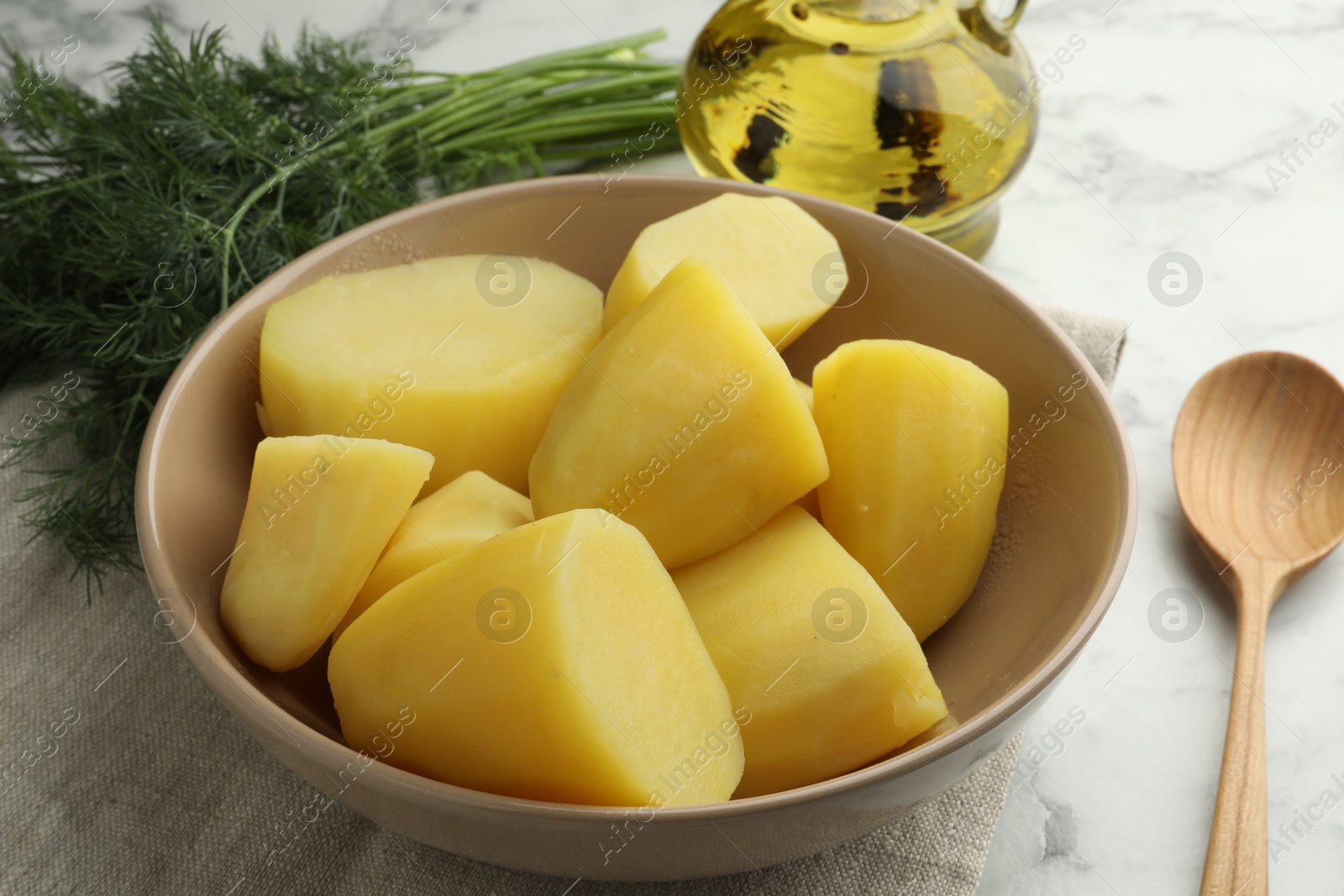 Photo of Boiled potatoes in bowl, dill, oil and spoon on white marble table