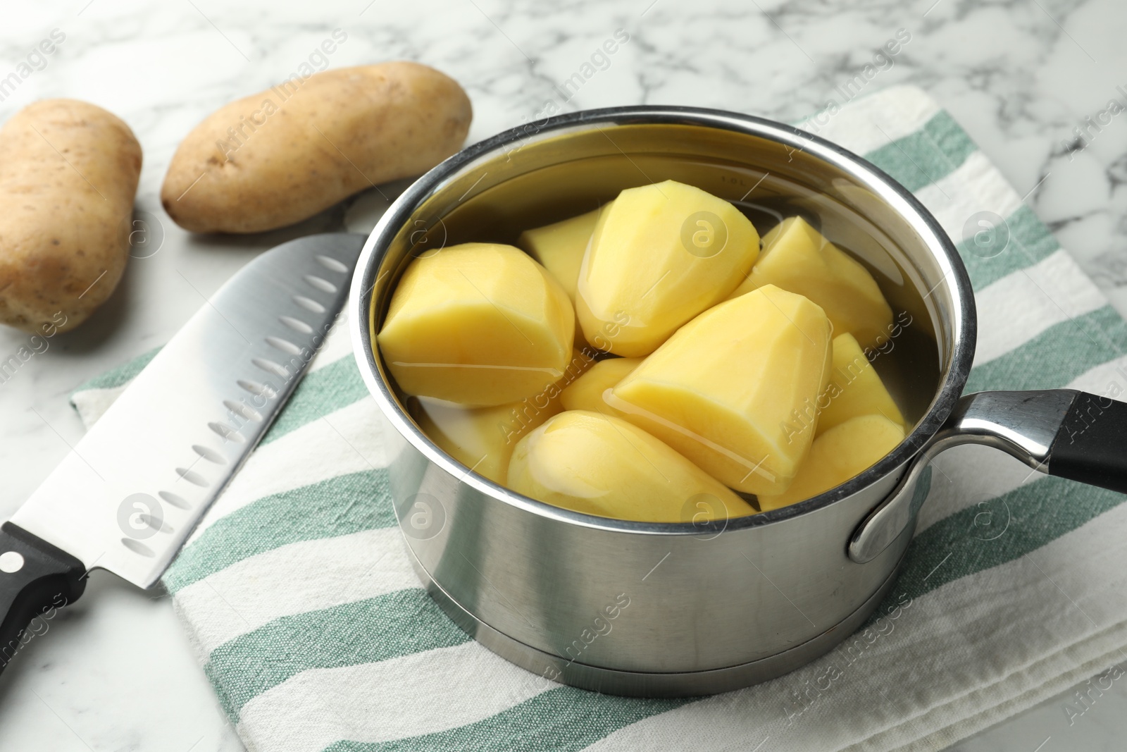 Photo of Raw potatoes in saucepan and knife on white marble table