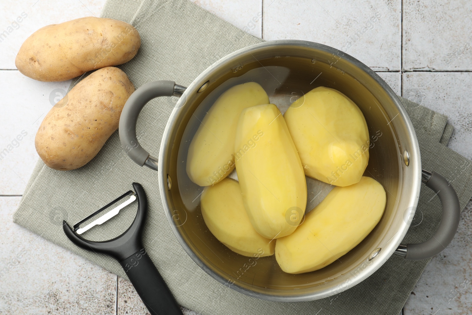 Photo of Raw potatoes in pot with water and peeler on tiled table, top view