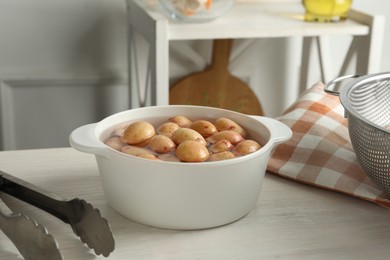 Photo of Raw potatoes in pot with water and tongs on white wooden table in kitchen