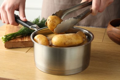 Photo of Woman putting raw potato into saucepan at wooden table, closeup