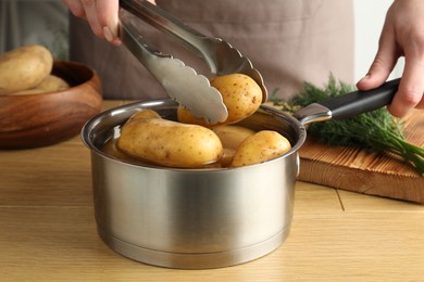 Woman putting raw potato into saucepan at wooden table, closeup