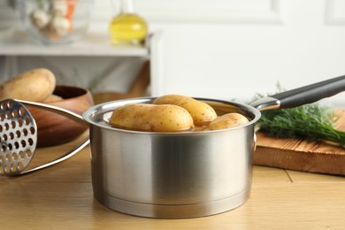 Photo of Raw potatoes in saucepan with water, pusher and dill on wooden table