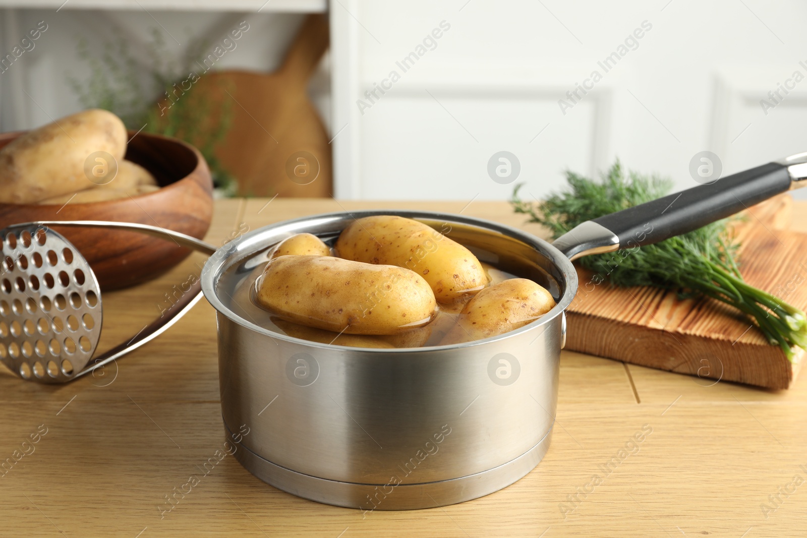 Photo of Raw potatoes in saucepan with water, pusher and dill on wooden table