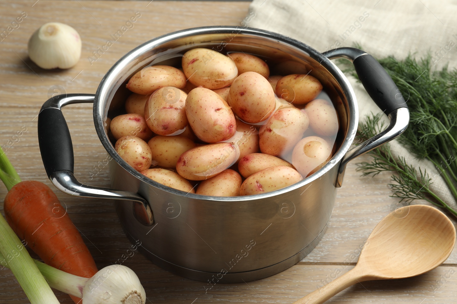 Photo of Raw potatoes in pot, green onion, carrot, dill and spoon on wooden table