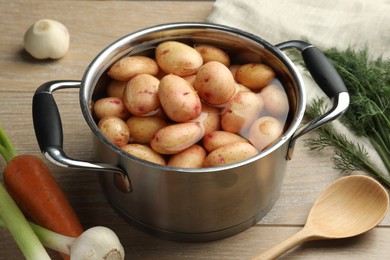 Photo of Raw potatoes in pot, green onion, carrot, dill and spoon on wooden table