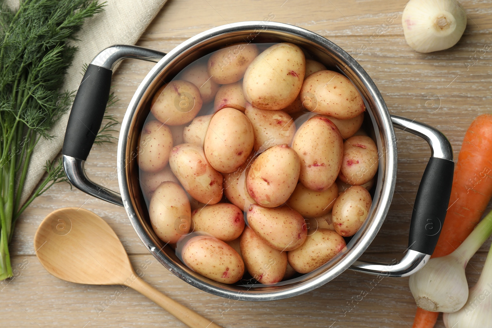 Photo of Raw potatoes in pot, green onion, carrot, dill and spoon on wooden table, top view