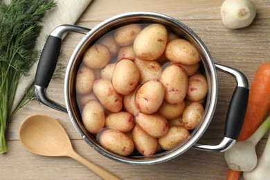 Raw potatoes in pot, green onion, carrot, dill and spoon on wooden table, top view