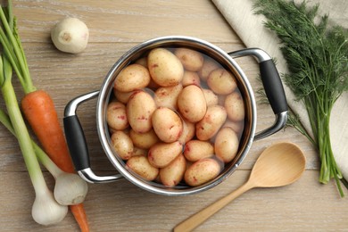 Photo of Raw potatoes in pot, green onion, carrot, dill and spoon on wooden table, top view