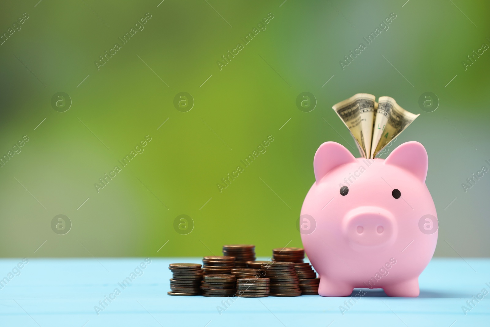 Photo of Pink piggy bank with dollar banknote and stacks of coins on light blue table against blurred background, space for text