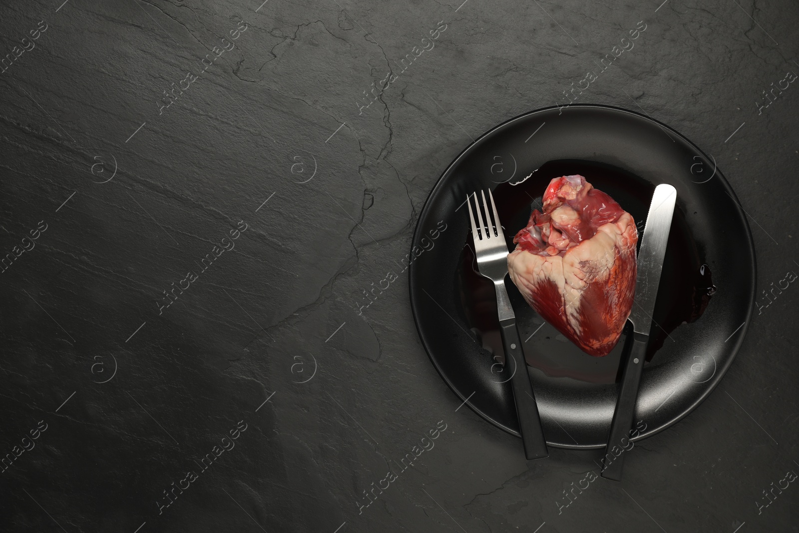 Photo of Raw heart, fork and knife on black table, top view. Space for text