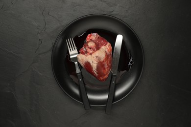 Photo of Raw heart, fork and knife on black table, top view