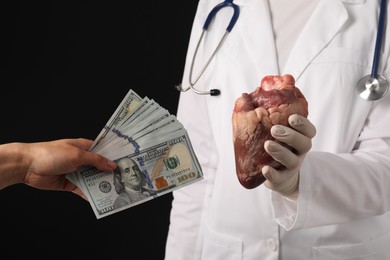 Photo of Patient giving dollar banknotes to doctor with heart on black background, closeup