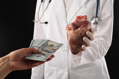 Patient giving dollar banknotes to doctor with heart on black background, closeup